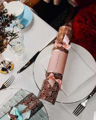 Two customised British Pullman Christmas crackers in chestnut and gold, with a blue and pink ribbon, rest on dinner plates.