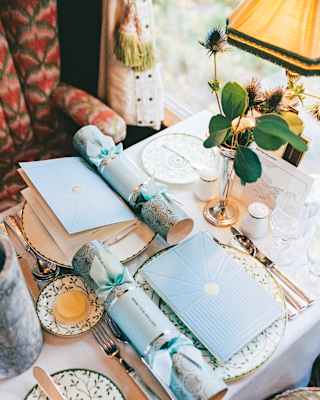 A Vera dining table awaits guests with a festive setting of crackers, tied with ribbons, and blue menus, seen from above.