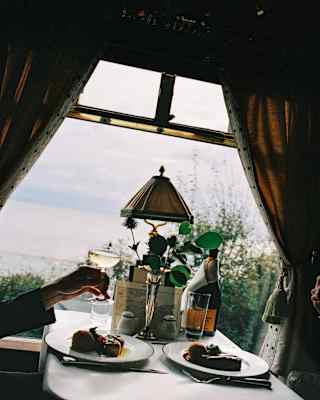 Angled eye-level image of a guest raising a Champagne glass at a table with two roast dinners, and curtains open to the view.