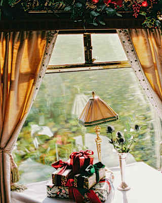 A pile of presents, wrapped with shiny paper and green and red bows rests on a window table below a festively decked pelmet.