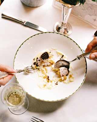 A couple at a table take a fork and spoon to share a dessert of mousse with chocolate and cream, seen close-up from above.