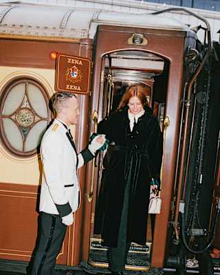A uniformed train steward holds the hand of a red-haired woman in a black coat as she steps down from the Zena carriage.
