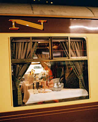 External view through the window of a warm-lit British Pullman carriage, glimpsing a table with a swan-shaped wine cooler.