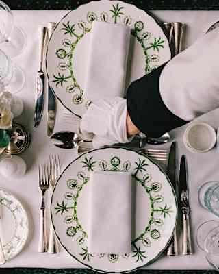 A waiter's gloved hand arranges cutlery between elegant Sultan's Garden crockery on a pretty dining table, seen from above.