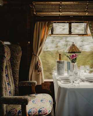 Light from a window brushes the seat of a dining chair, in shades of taupe, blue and purple, next to a table laid for dinner.