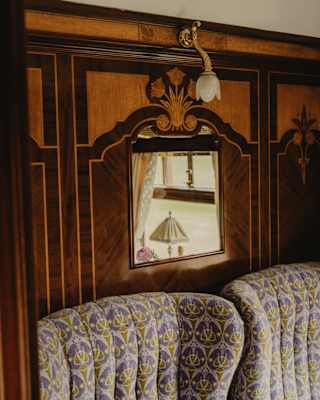 Above two grey and purple fabric seats, a mirror set into a marquetry panel with a rose motif reflects a window and lamp.