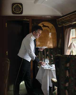 A waiter sets a table in an art deco carriage. Divine marquetry on the wall behind him shows a gazelle leaping a gold hillock