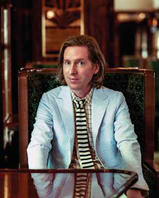 Wes Anderson smiles quizzically, dressed in a pale blue seersucker blazer, yellow and navy striped tie and yellow plaid shirt