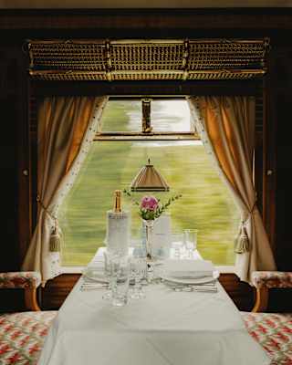 A view flies by a window table, laid for two, with Champagne and a bud vase, flanked by Zena's pink and grey-patterned seats.