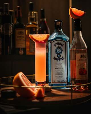 A blue Bombay Sapphire bottle sits between a luminous tall orange drink and a liquor bottle, both topped with citrus slices.