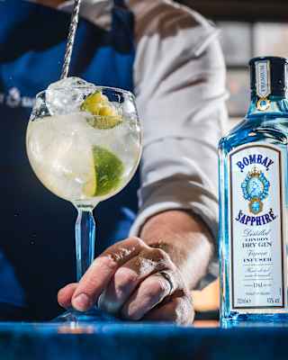A barman, seen from the neck down, uses a swizzle to stir a lime-garnished glass of gin, alongside a Bombay Sapphire bottle.