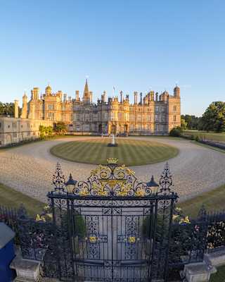 High-angle view of the large Burleigh House estate, stone glowing gold in late sun, seen over the wrought iron gates.