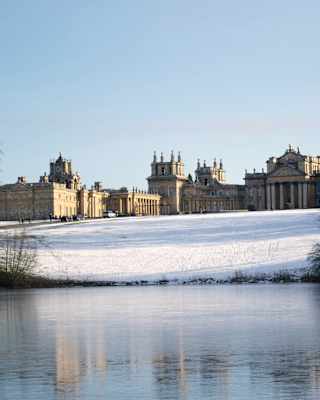 Blenheim Palace, in winter sun, sits atop a gentle sloping field covered with snow, which reflects on the icy lake water below.