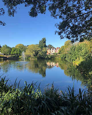 Bletchley Park stands grand beneath a clear blue sky, in a view across the lake that captures the tree-filled verdant estate.