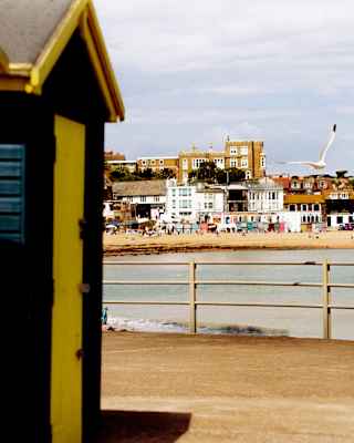 A seagull cruises over the water in a view across Broadstairs harbour, part-obscured by two yellow and black beach huts.