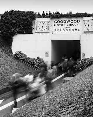 Blurred monochrome shot of crowds running through an underpass below a bridge with two clocks flanking a Goodwood sign.