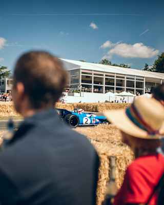 A blue car zips past on a track with haybale barriers, lined with spectators at the Goodwood track, seen through crowds.