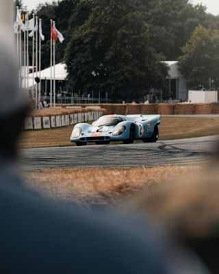 A light blue Porsche nine-one-seven race-car speeds on the track at Goodwood Festival of Speed, glimpsed through the crowd.