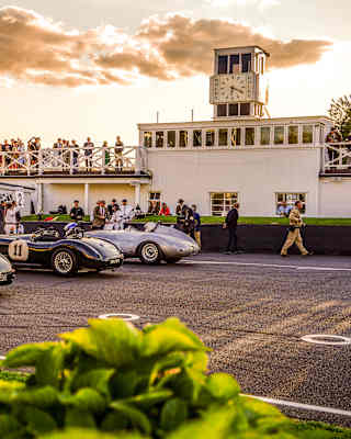 Saturated image of the track where three classic cars wait in line, and crowds mill at the side or watch from a roof terrace.