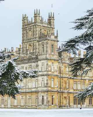 An impressive view of Highclere Castle with towering turrets and intricate roofscape, flanked by enormous snowy fir trees.