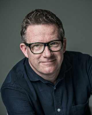 A man with short brown hair, wearing black glasses and a dark button-up shirt, smiles softly whilst sitting against a plain, dark background.