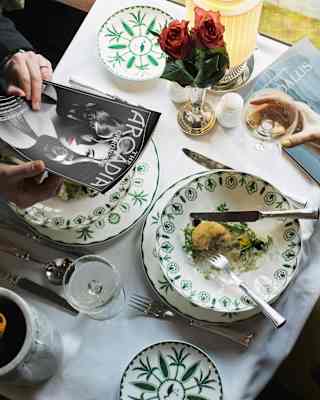 Looking down on a dining table, where a man peruses a magazine and a woman takes a pause mid-way through a starter to drink.