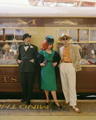 A woman with red curly hair in a green dress and hat leans against the stationed train flanked by two men, all in costume.