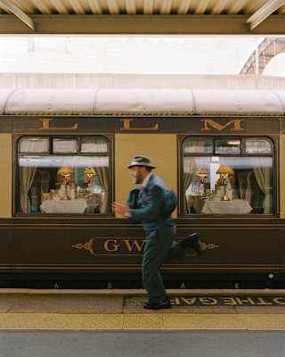 A man, dressed in a blue nineteen-thirties suit and hat runs in a blur on the platform in front of the stationed Gwen car.