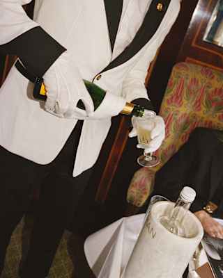 A waiter in a black and white tux pours Champagne from a bottle into a tulip glass, in an angled image taken in the Gwen car.