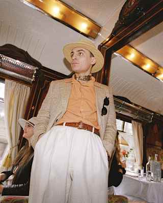 A man in white chinos, peach shirt and beige jacket and hat, stands with hands in pockets, seen in a carriage at a low-angle.