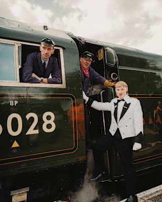 A steward stands on a platform, leaning on the doorframe of the train where one driver stands as another looks from a window.