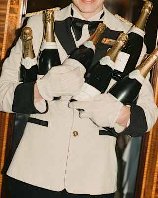 A British Pullman steward in a white jacket with black detailing holds six bottles of Champagne against his chest with gloved hands.