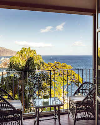 A balcony with wicker chairs and a drinks table takes in the blues of ocean and coastline views, over the fringes of a palm.