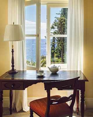 An elegant antique wooden desk with a lamp and a pot of tea by an open window with views of the botanical gardens and sea.