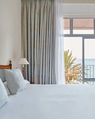 A large window flanked by drapes brings sublime sea views into a room with a white armchair and bed with two blue cushions.