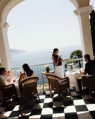 Tilted image of two couples, one woman standing, as they take afternoon tea on the ocean-view, monochrome-tiled tea terrace.