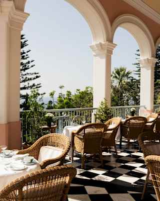 View of the Tea Terrace with laid tables, wicker chairs, a monochrome-tiled floor and wide arches framing heavenly sea views.