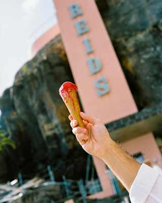 A man's hand holds up a cone of dripping raspberry-pink gelato in front of the Reid's Palace pink cliff-face column.