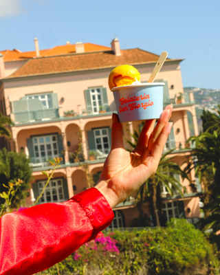 An arm in a red sleeve holds up a blue tub with a boule of mango-hued ice-cream in front of Reid's Palace hotel and gardens.