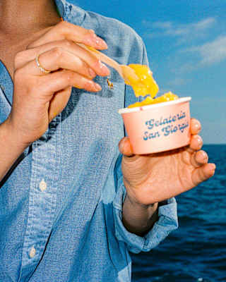 A woman in a blue shirt, seen from the neck down, scoops mango-hued gelato from a Gelateria San Giorgio pot, by the ocean.