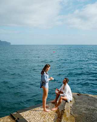 A woman, standing in bare legs and a blue shirt chats with a man in a white top seated on the swimming platform by the ocean.