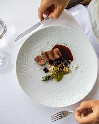 A diner holds a knife and fork by a plate of seared pork and asparagus at a table with red wine and water, seen from above.