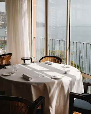 A round window table with four chairs, water glasses and rolled napkins catches the sun and sea views at William Restaurant.