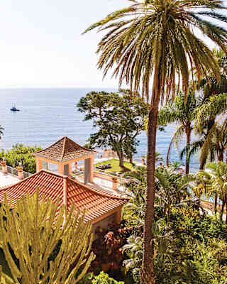 View of azure ocean, seen over the terracotta-tiled gazebo in emerald-green gardens with palms, cacti and empress trees.