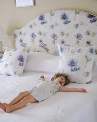 A smiling young child in short dungarees lies on their back with arms spread on a fresh white bed with floral cushions.
