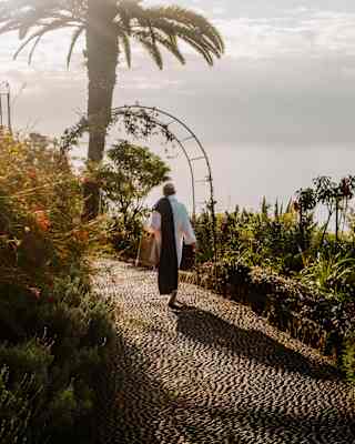 A guest in a white robe with a towel on one shoulder casts a shadow behind him as he walks on a garden path with sea views.