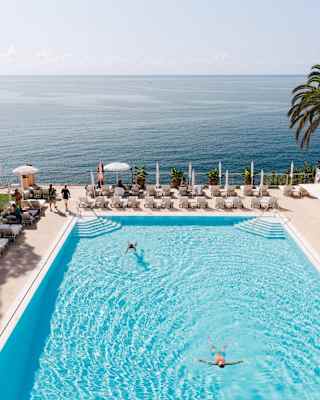 Two guests swim in the large, sparkling blue pool, which offers uninterrupted views of deeper ocean hues, seen from above.