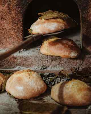 Close-up of three round breads by a brick oven as a cook, off-camera, uses a peel to lift another from the oven opening.