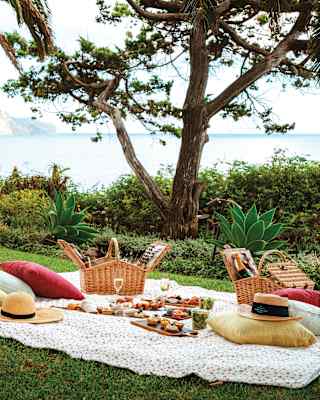 In a corner of lawn overlooking garden beds and the ocean, a quilt covered with food, hampers and cushions awaits picnickers.