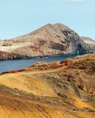 View over a rocky ridge with gold grasses to a deep blue inlet where four small boats are anchored by Ponta de São Lourenço.